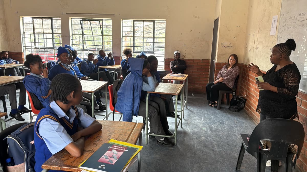 A teacher speaking to learners in school uniform seated at wooden desks in a classroom, while a volunteer looks on