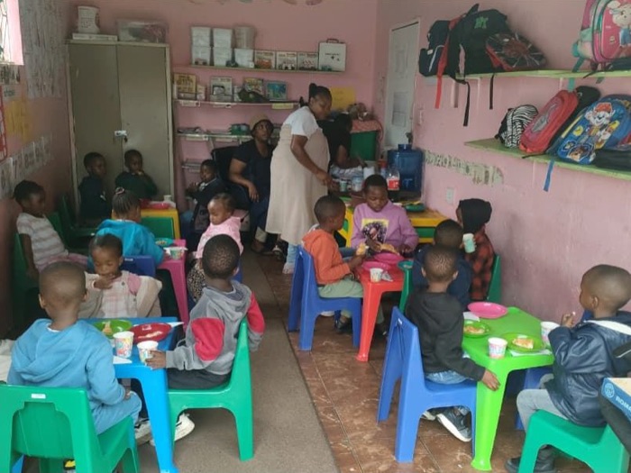 Children sitting at small tables sharing a meal in a preschool classroom while staff look after them